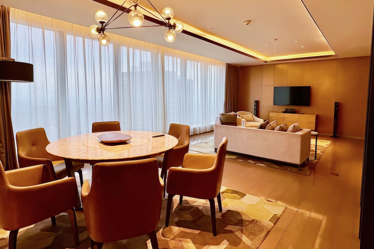 A modern living room with a round dining table, comfortable seating, a sofa, a flat-screen TV, and a large window with curtains.