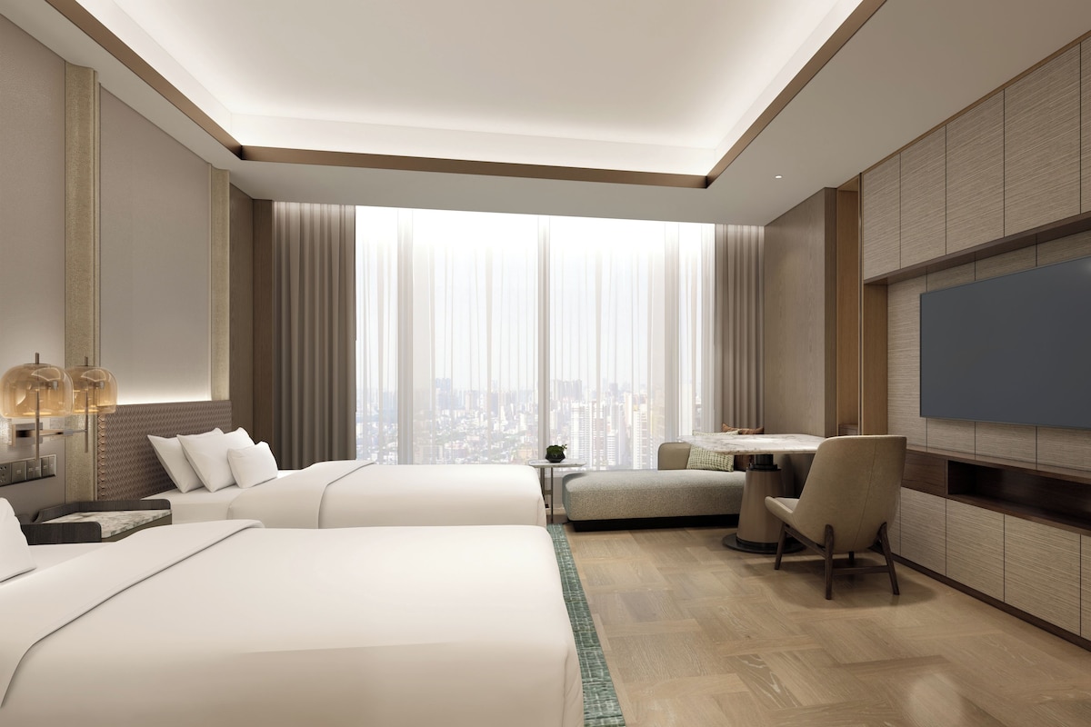 A modern hotel room with a large bed, a desk, a chair, a TV, and a city view.