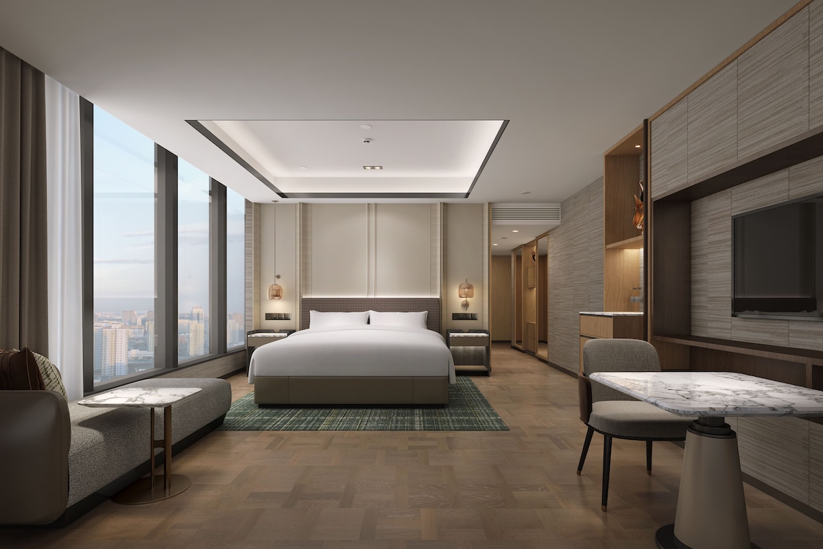 A modern hotel room with a large bed, a seating area, a dining table, and a TV.