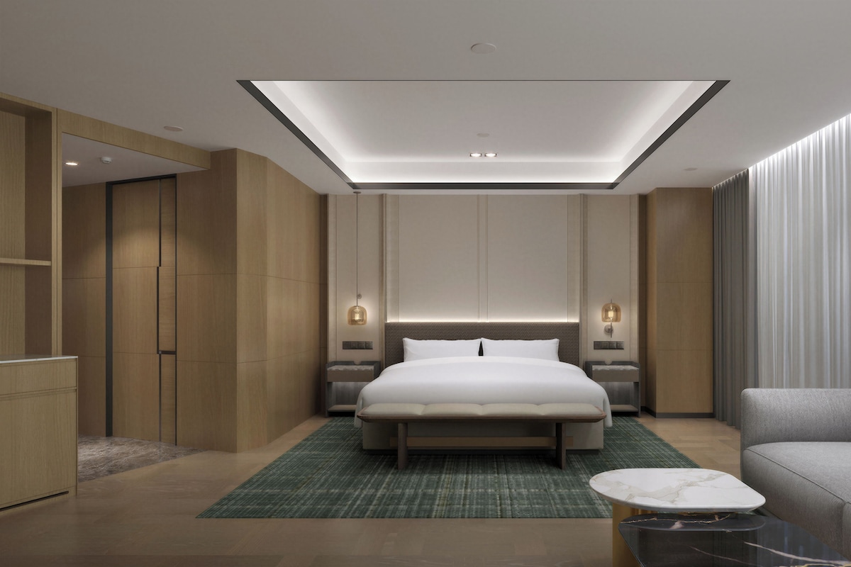 A modern hotel room with a large bed, bedside tables, a sofa, a small table, and a ceiling skylight.