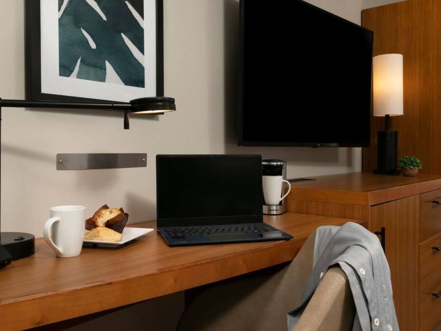 A hotel room with a wooden desk, a laptop, a coffee maker, a telephone, a framed picture, a television, and a cabinet with drawers.