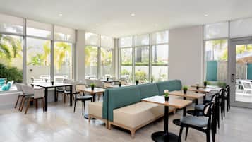 A modern indoor dining area with large windows, a green cushioned bench, and multiple tables and chairs.