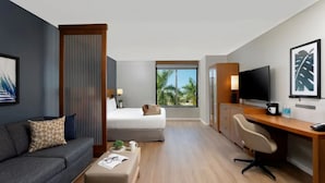 Habitación de hotel con cama, escritorio, televisor, sofá y ventana con vista a palmeras.
