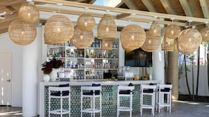 Zona de bar amb llums penjants de vimet, tamborets blancs i diverses ampolles a les prestatgeries.