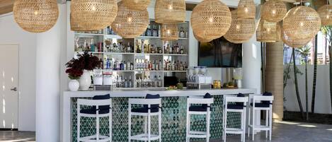 A bar area with wicker pendant lights, white bar stools, and a variety of bottles on shelves.
