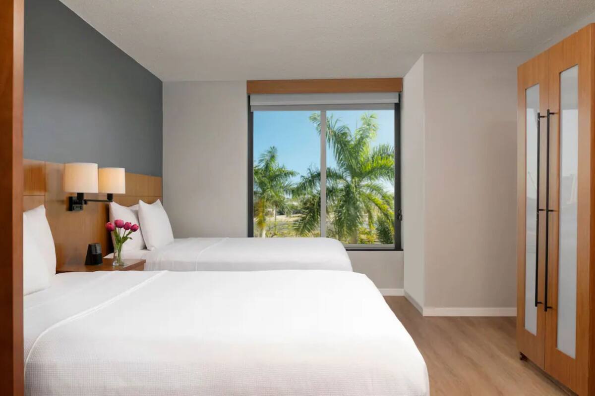 A hotel room with two beds, a large window with a view of palm trees, a wooden wardrobe, and a nightstand with a lamp and flowers.