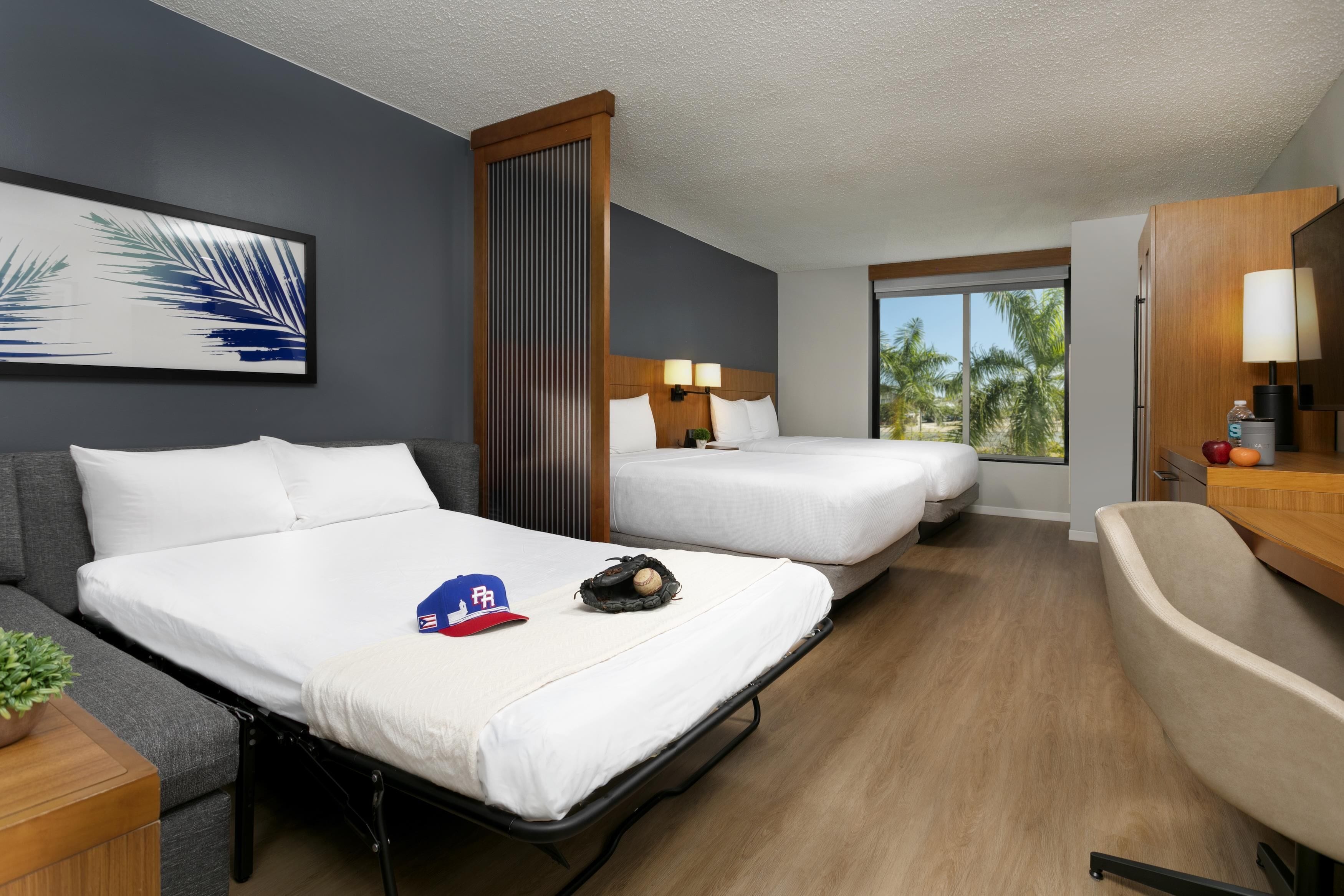A hotel room with two beds, a sofa, a desk, a lamp, a window with a view of palm trees, and a framed picture on the wall.