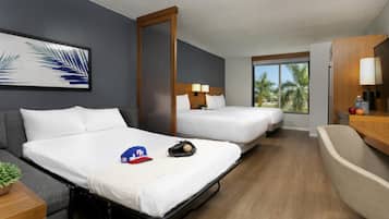 A hotel room with two beds, a sofa, a desk, a lamp, a window with a view of palm trees, and a framed picture on the wall.