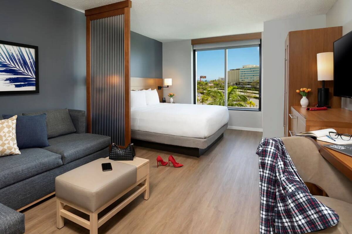 A hotel room with a bed, a sofa, a desk with a laptop, a TV, and a window with a city view.