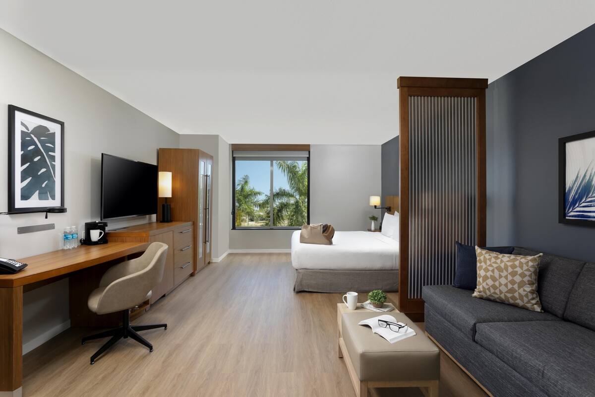 A modern hotel room with a wooden desk, a bed, a sofa, and a large window with a view of palm trees.