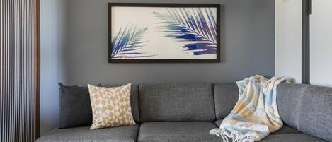 A grey sofa with patterned cushions, a framed leaf artwork on the wall, a small plant on a side table, and a lamp.