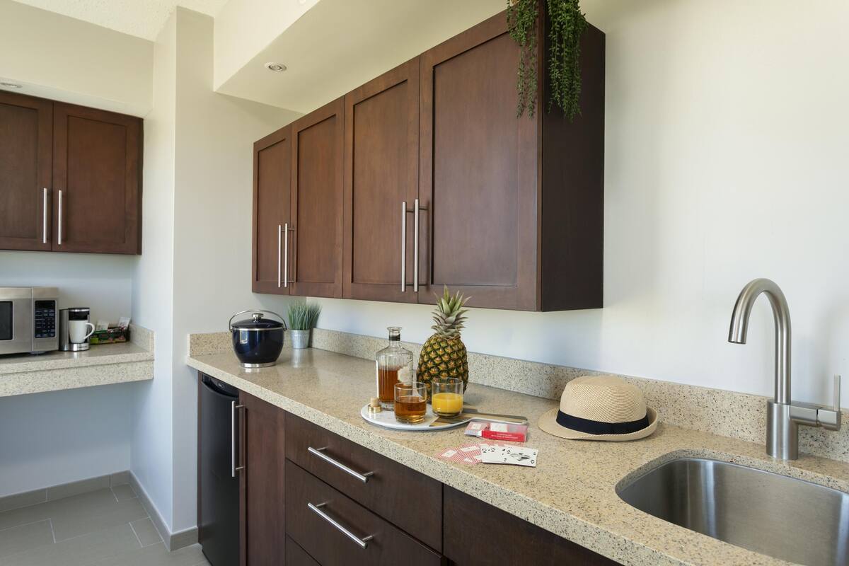 A kitchen with wooden cabinets, a stainless steel sink, a microwave, and a countertop with a pineapple, a hat, and a bottle.