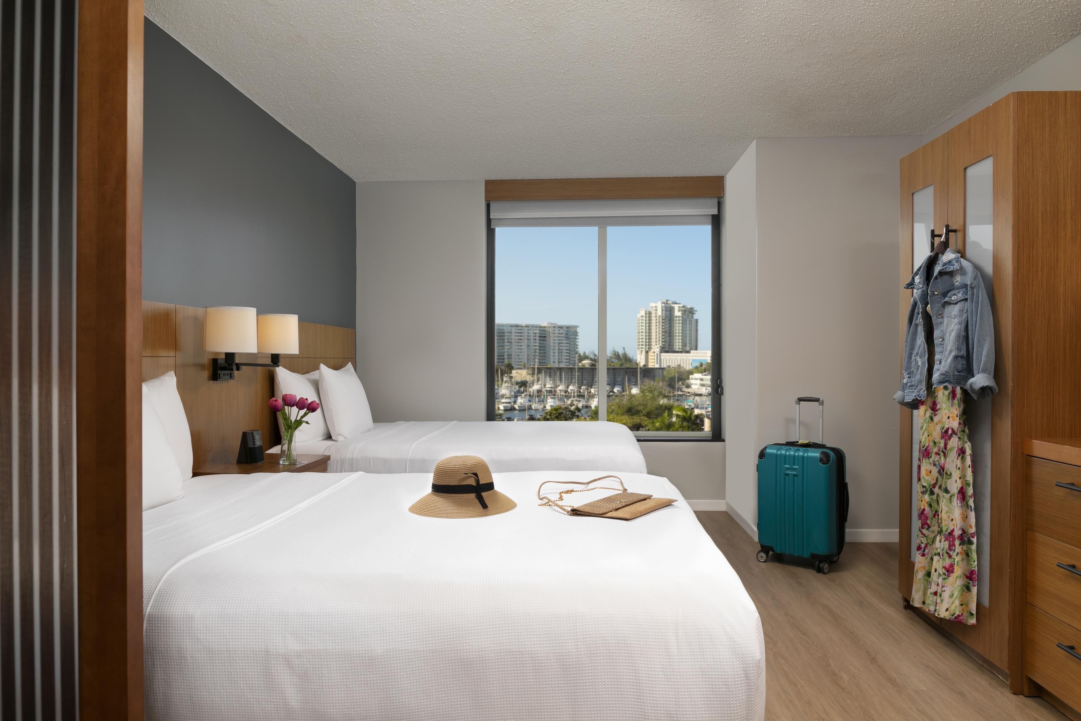 A hotel room with two beds, a large window with a city view, a suitcase, and a jacket hanging on the wall.