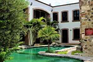 A courtyard with a pool, palm trees, and a building with arched windows and a stone wall. - Hotel Hacienda San Cristobal (San Francisco del Rincon)