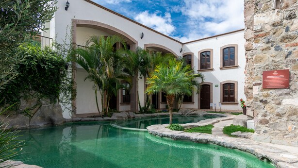 A pool area with a clear blue pool, surrounded by a white building with arches and a stone wall.