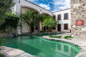 Un área de piscina con una piscina azul cristalina, rodeada por un edificio blanco con arcos y un muro de piedra.