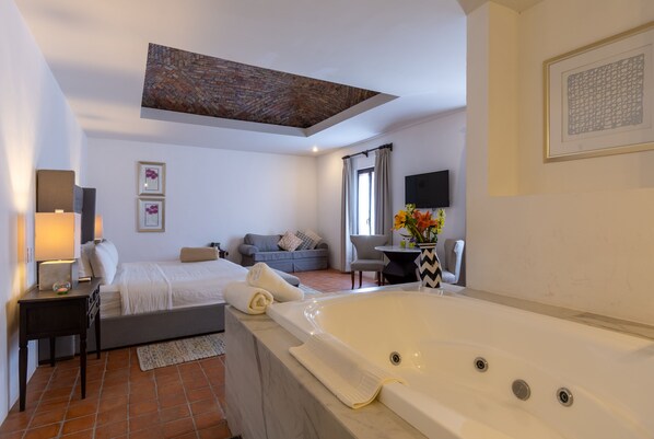 A modern hotel room with a large bathtub, a sofa, a dining table, and a TV. - Hotel Hacienda San Cristobal (San Francisco del Rincon)