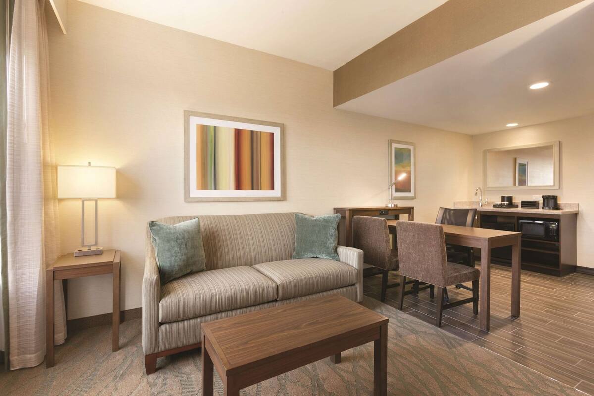 Une chambre d’hôtel comprenant un canapé, une table basse, un bureau avec des chaises, un coin cuisine et un tableau abstrait encadré accroché au mur.