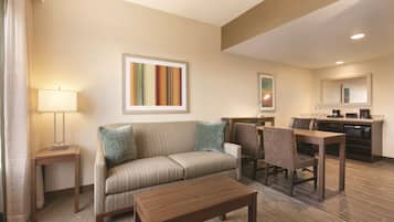 Une chambre d’hôtel comprenant un canapé, une table basse, une table à manger avec des chaises, un coin cuisine et une salle de bain avec un miroir.