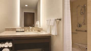Une salle de bain d’hôtel équipée d’un lavabo, d’un miroir, d’un porte-serviettes, d’une douche et d’un siège de douche en verre.