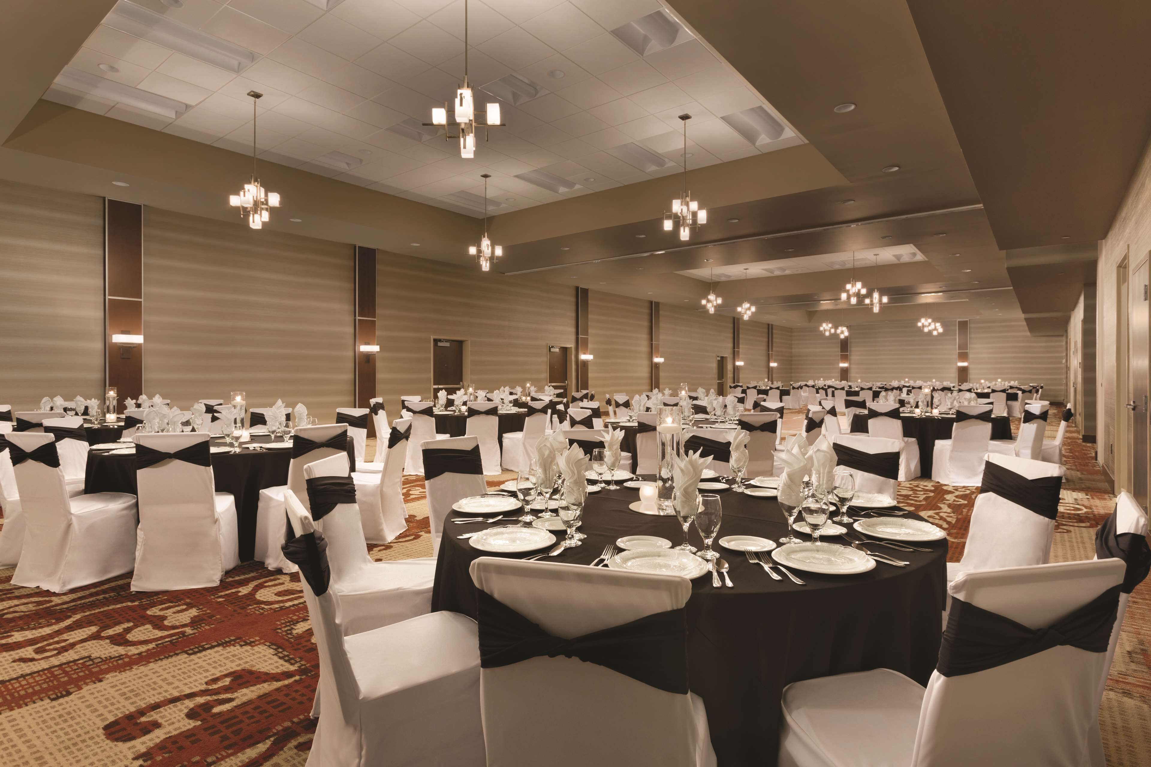 A banquet hall with round tables set for dining, each with white tablecloths and black sashes, and chairs with white covers and black sashes.