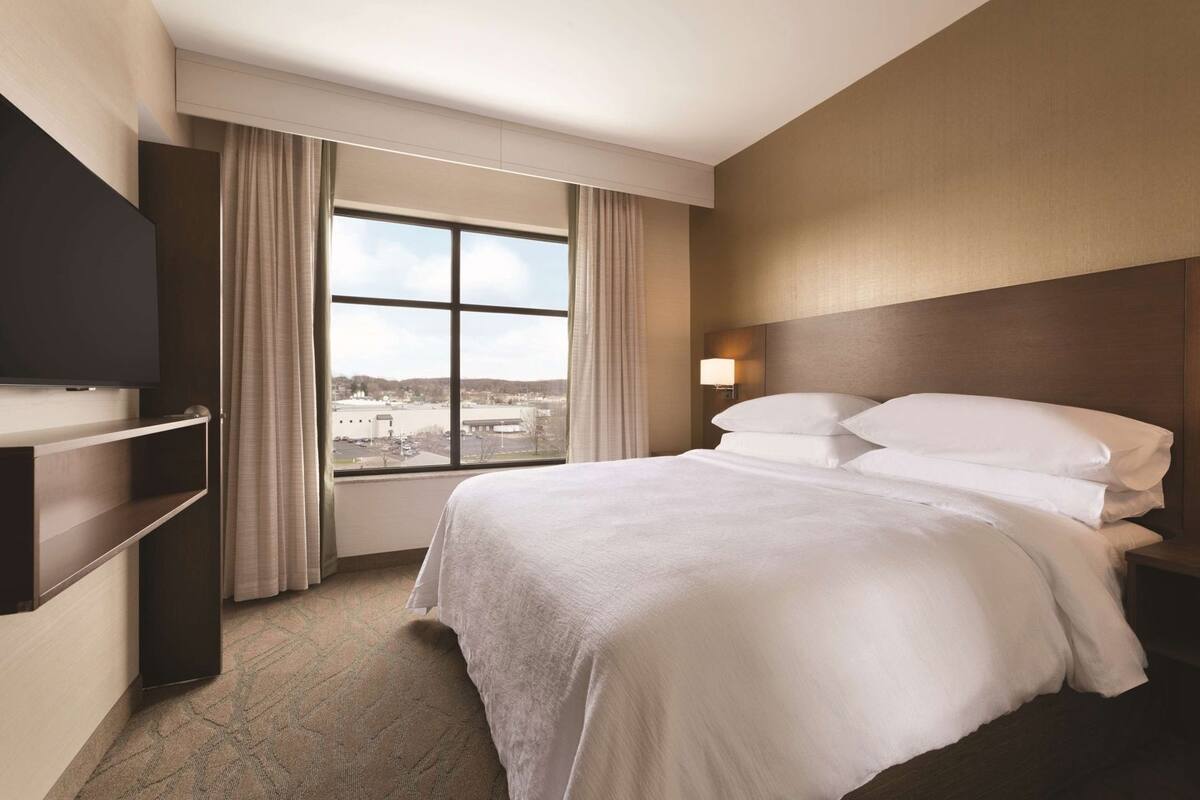 Une chambre d’hôtel avec un grand lit, un téléviseur fixé au mur, une fenêtre avec des rideaux et une table de chevet avec une lampe.