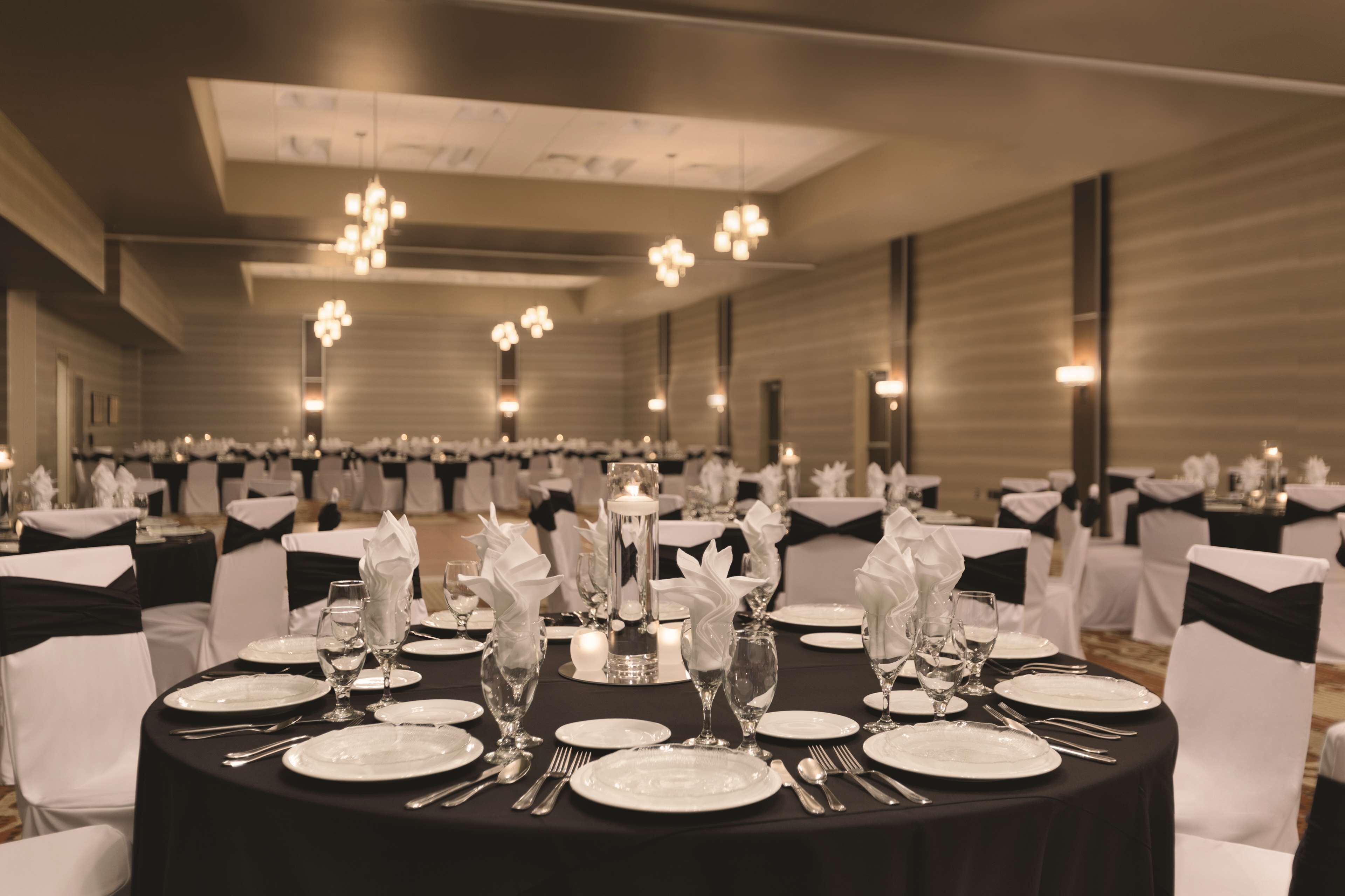 Un salón de banquetes con mesas redondas dispuestas para un evento formal, con manteles negros, sillas blancas y centros de mesa elegantes.