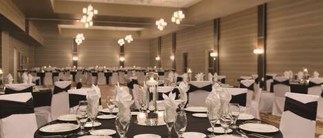 Un salón de banquetes con mesas redondas dispuestas para un evento formal, con manteles negros, sillas blancas y centros de mesa elegantes.
