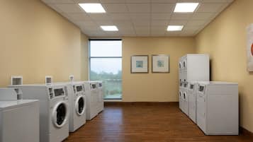 A laundry room with multiple washing machines and dryers, wooden flooring, and a large window providing natural light.