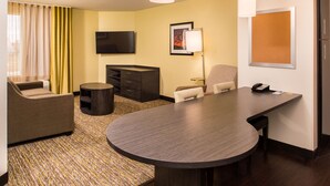 Una habitación de hotel con una mesa redonda grande, un sofá, un televisor, un escritorio con teléfono y una lámpara.