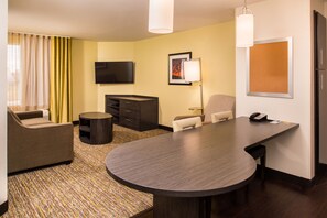 A hotel room with a large round table, a sofa, a TV, a desk with a phone, and a lamp. - Candlewood Suites Carlsbad South by IHG (Carlsbad)