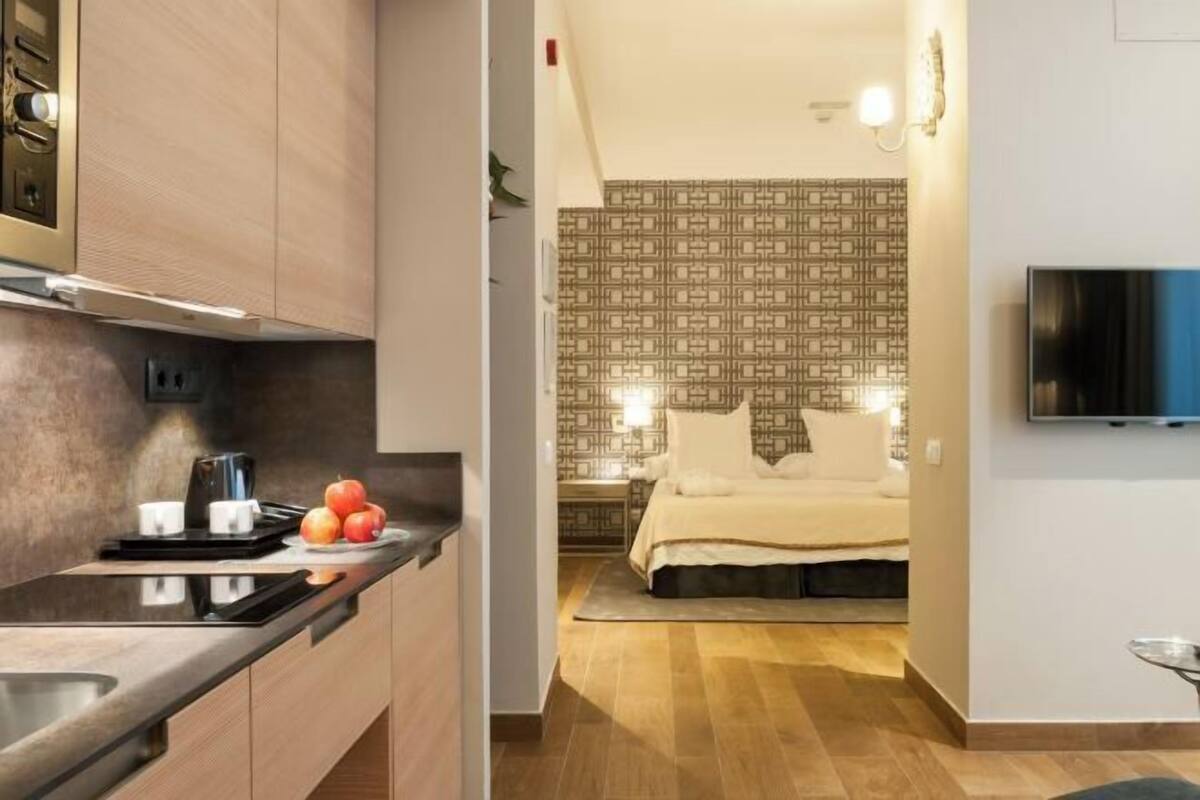 A modern hotel room with a kitchenette, a bed with a patterned headboard, and a flat-screen TV mounted on the wall.