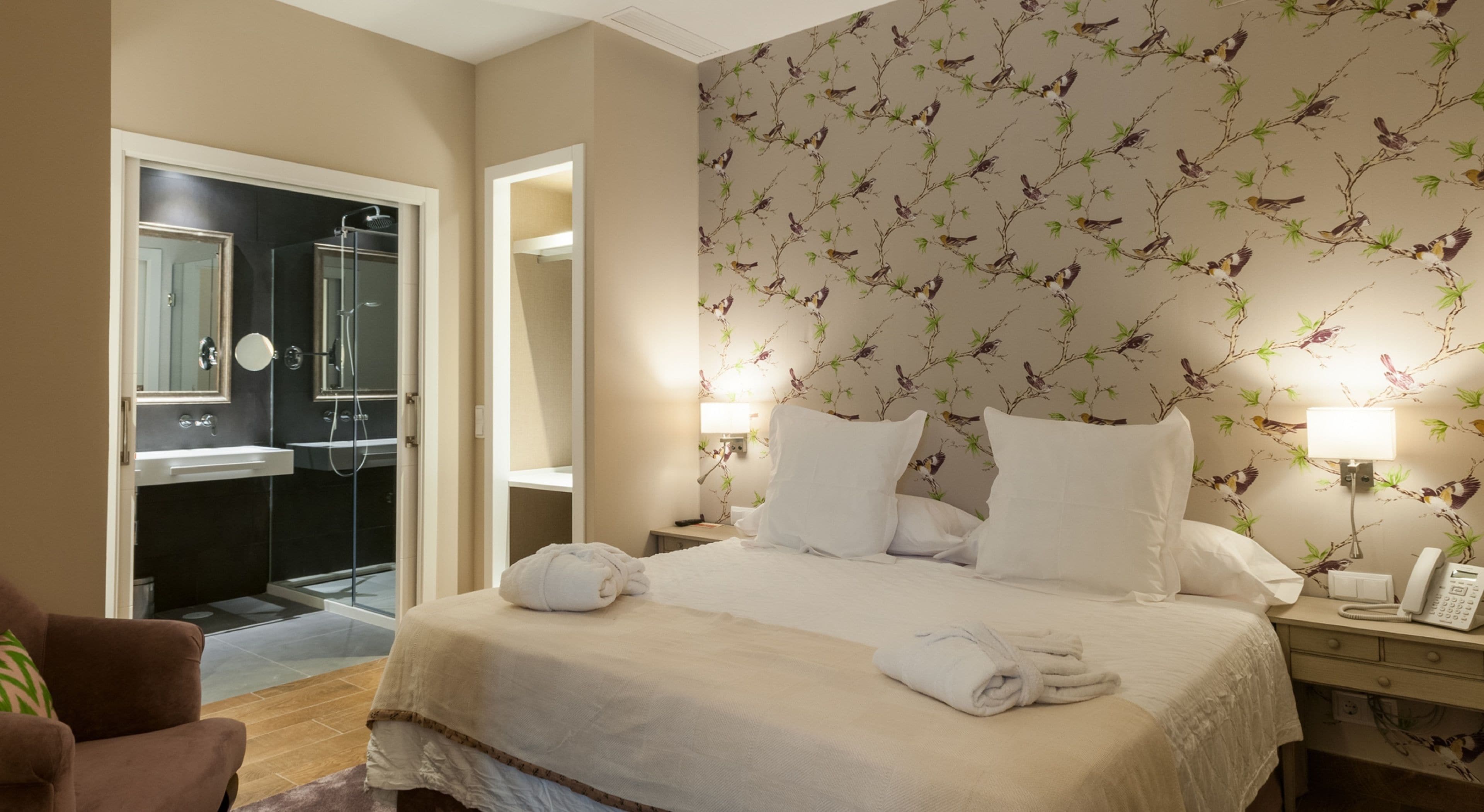 A hotel room with a bed, two bedside lamps, a nightstand with a phone, a bathroom with a shower and sink, and a door leading to another room.