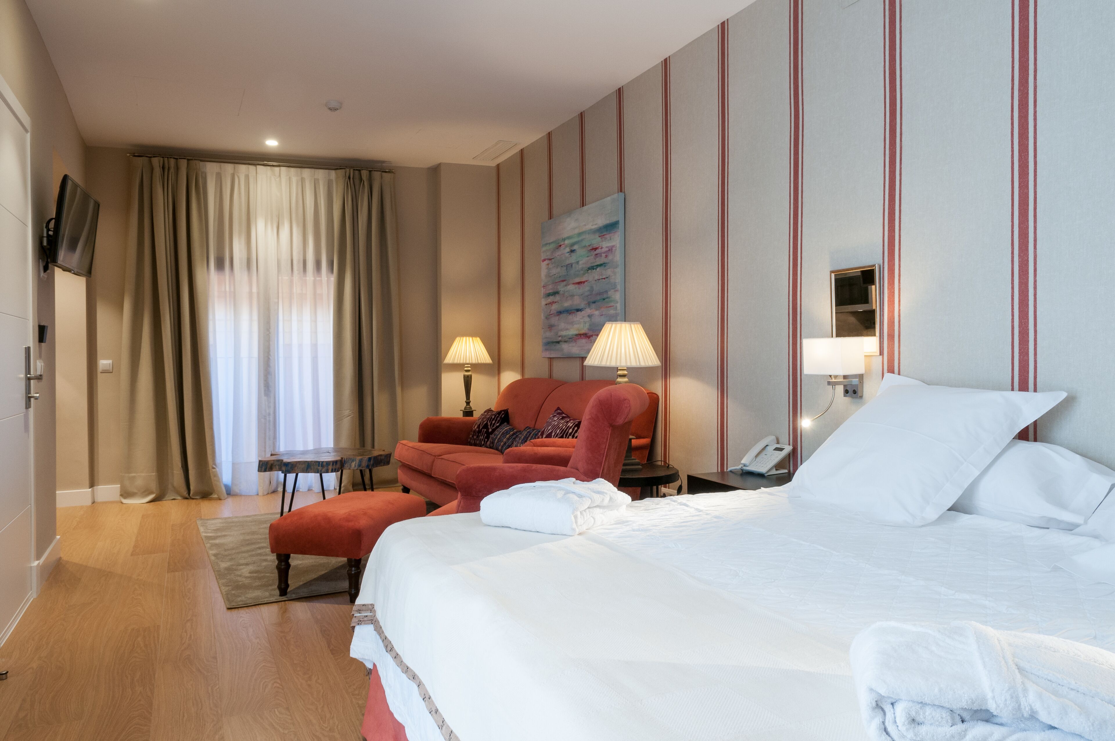 A hotel room with a large bed, a red armchair, a small table, and a TV mounted on the wall.