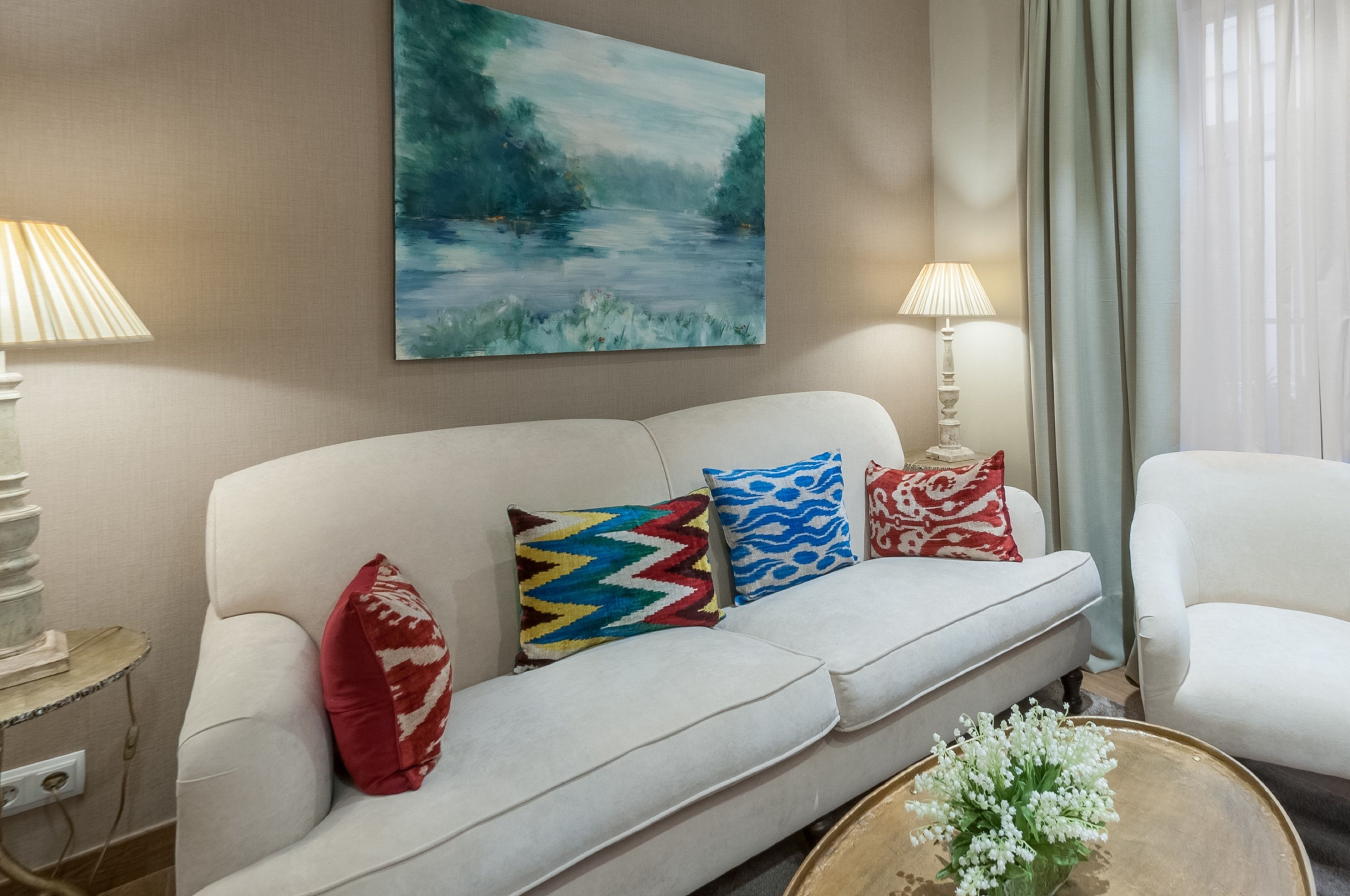 Un salon avec un canapé blanc, des coussins colorés, un tableau accroché au mur et une lampe posée sur une table d’appoint.