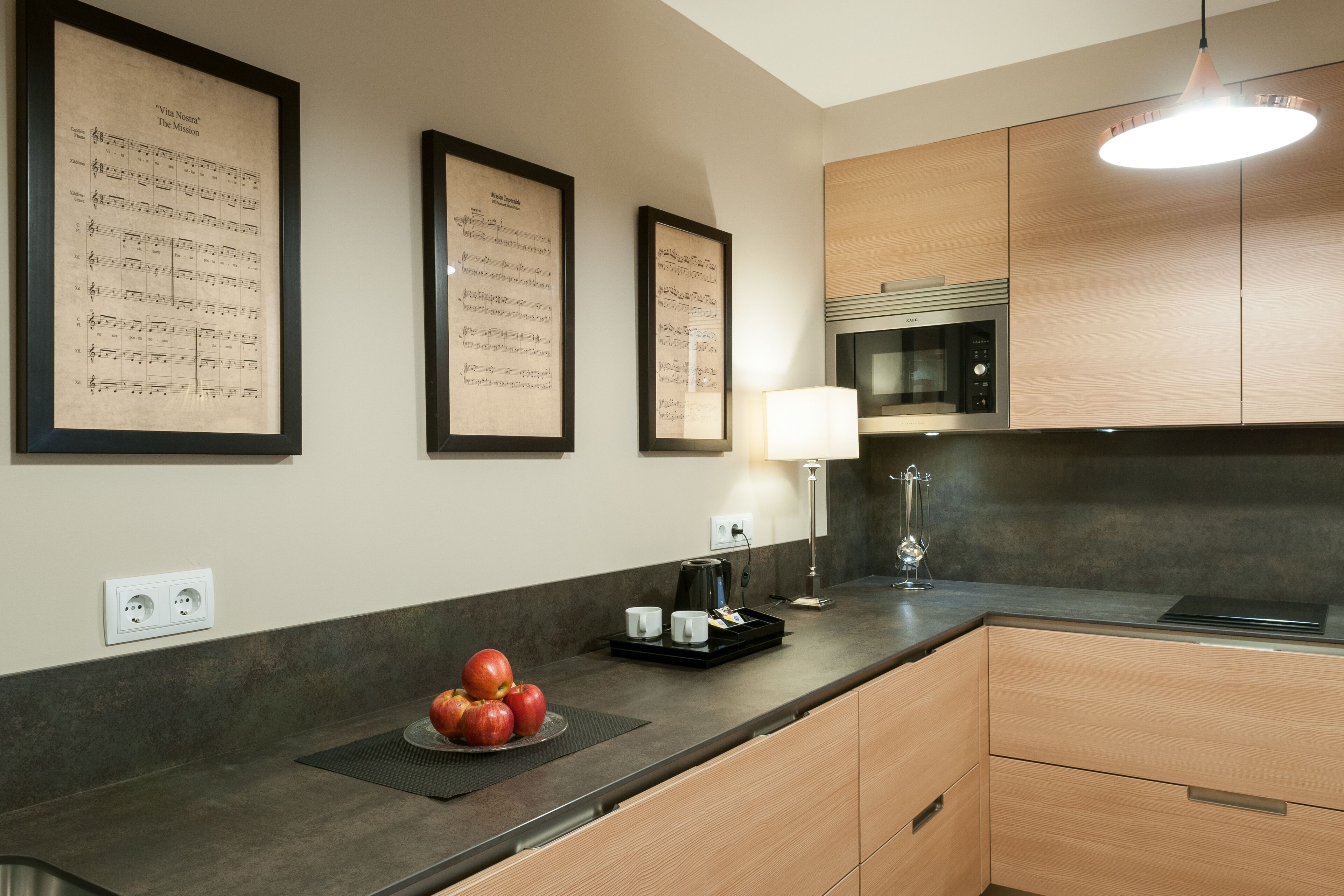 A modern kitchen with wooden cabinets, a black countertop, and framed sheet music on the wall.