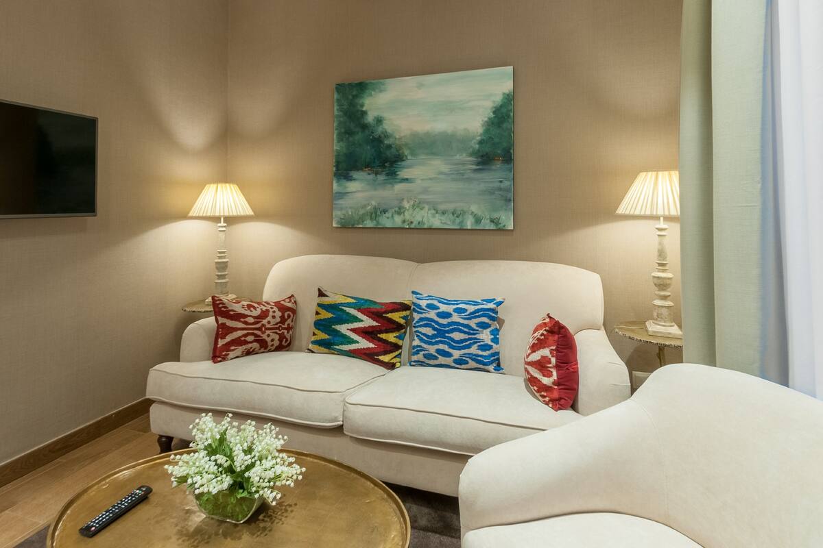 A living room with a white sofa, colorful pillows, a painting, two lamps, a remote control, and a vase with flowers on a coffee table.