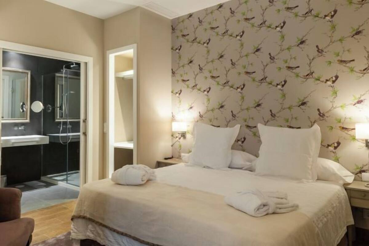 A hotel room with a bed, two bedside lamps, a nightstand, a bathroom with a shower, and a mirror.