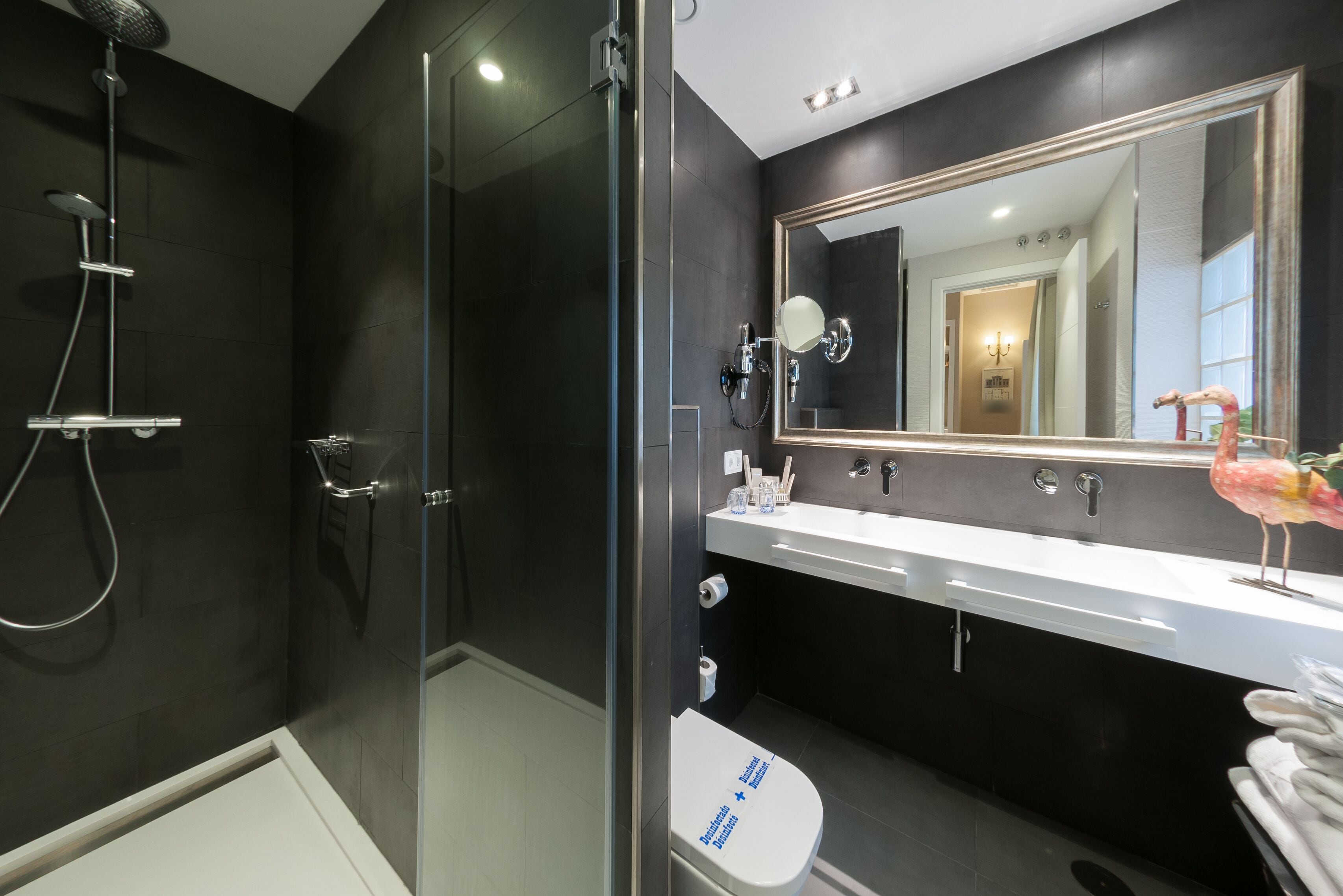 A modern bathroom with a glass-enclosed shower, a white toilet, and a large mirror above a vanity with a white countertop.