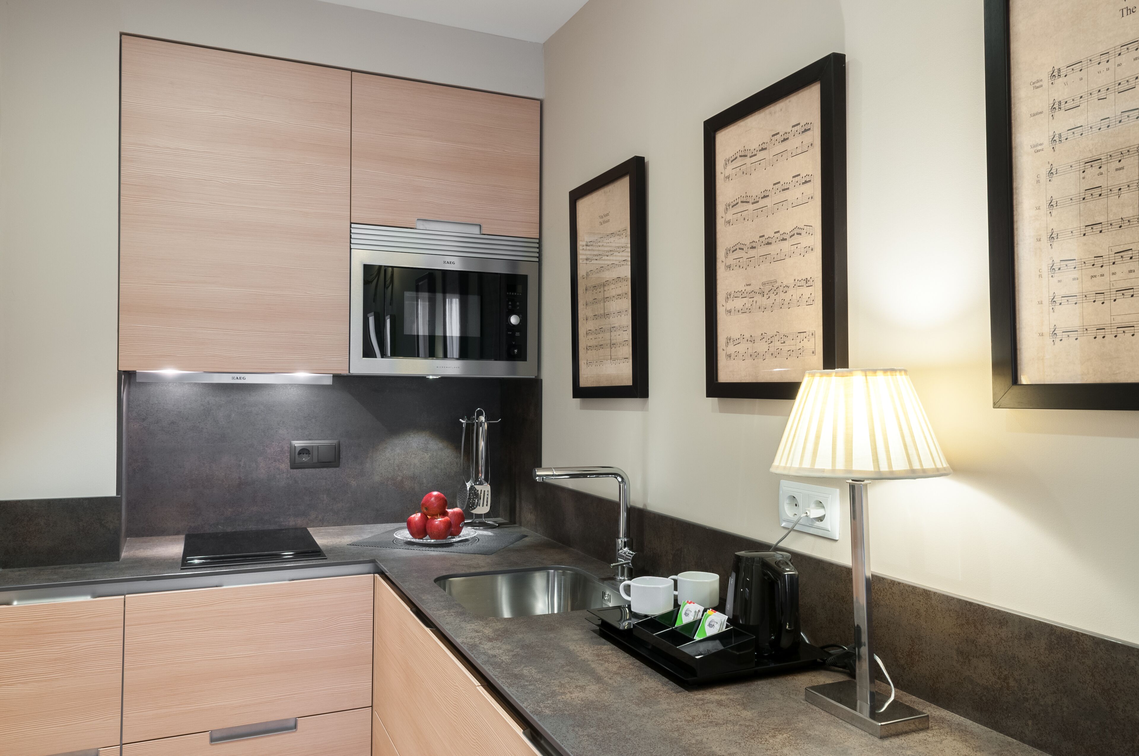 A modern kitchen with a built-in microwave, stainless steel sink, and a kettle. There are framed musical sheets on the wall and a lamp on the counter.