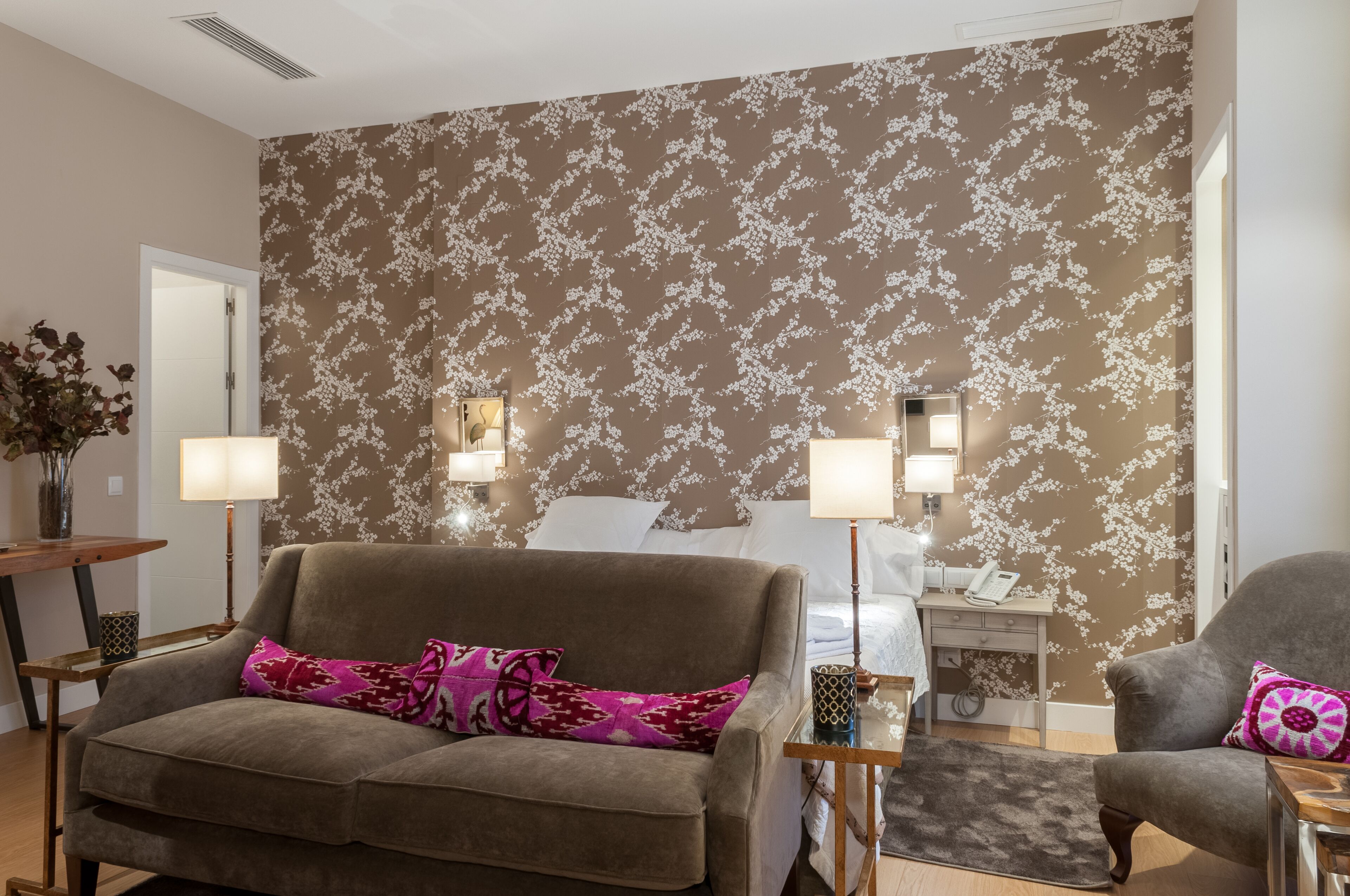 Habitación de hotel con papel tapiz floral, un sofá, una silla, una mesita de noche y una lámpara.