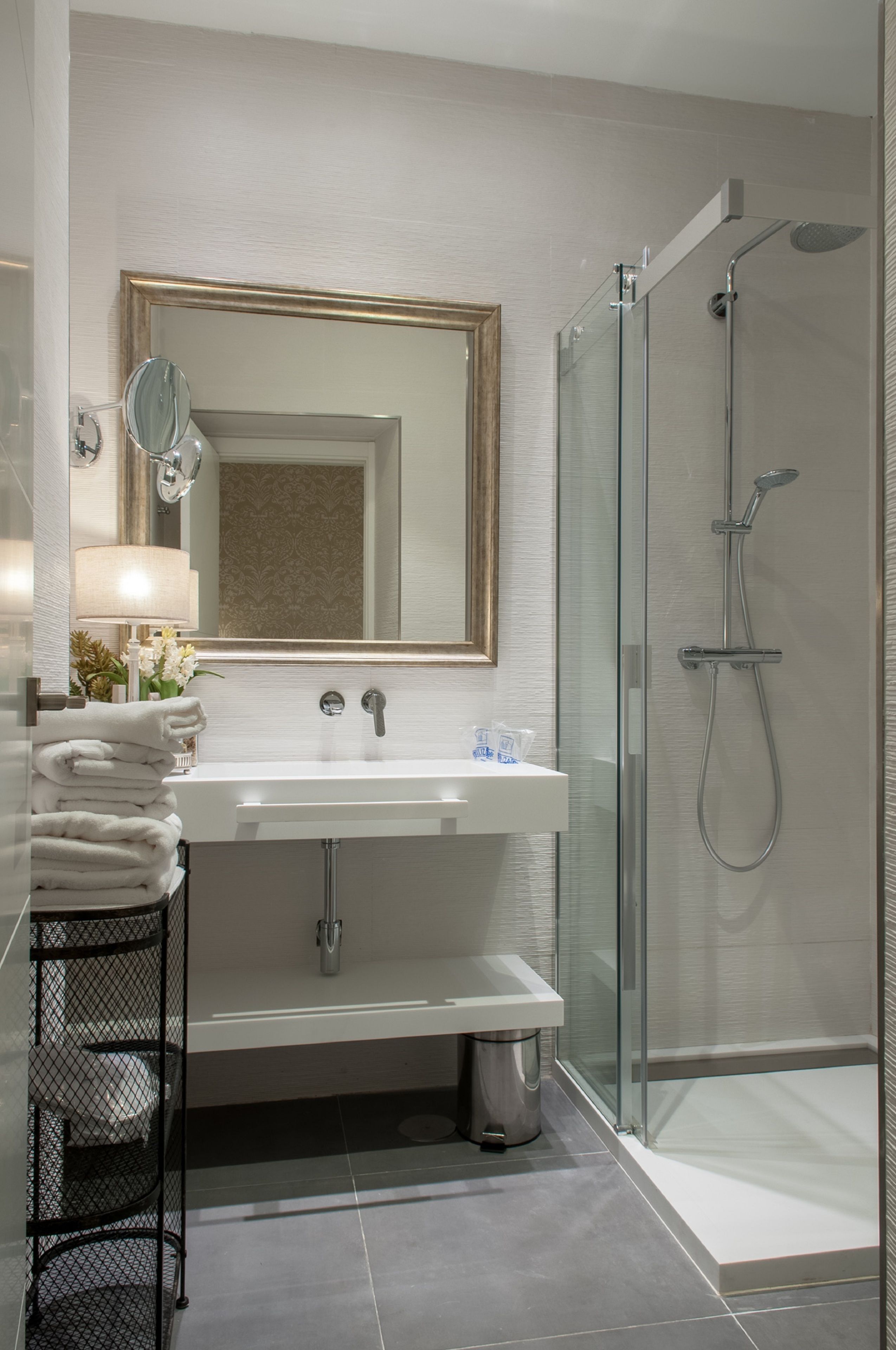 A modern bathroom with a glass-enclosed shower, a white sink with a mirror above it, and a black metal shelf with towels.