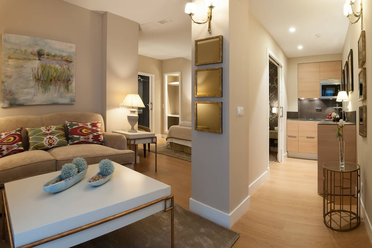 A modern living room with a sofa, coffee table, and decorative items. A hallway leads to a kitchen area with cabinets and a microwave.