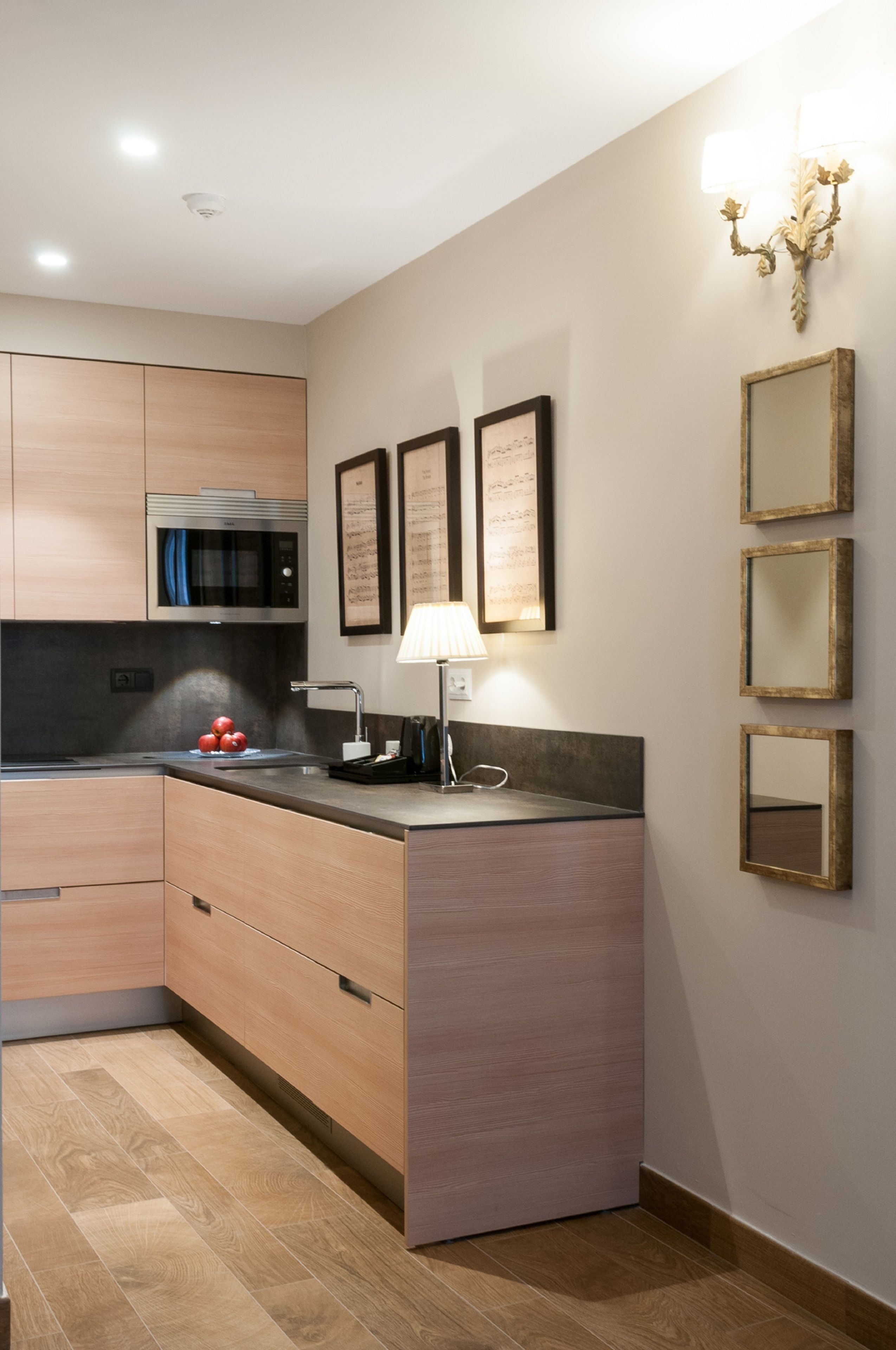 A modern kitchen with wooden cabinets, a built-in microwave, a sink, and a countertop. There are framed pictures on the wall and a chandelier.