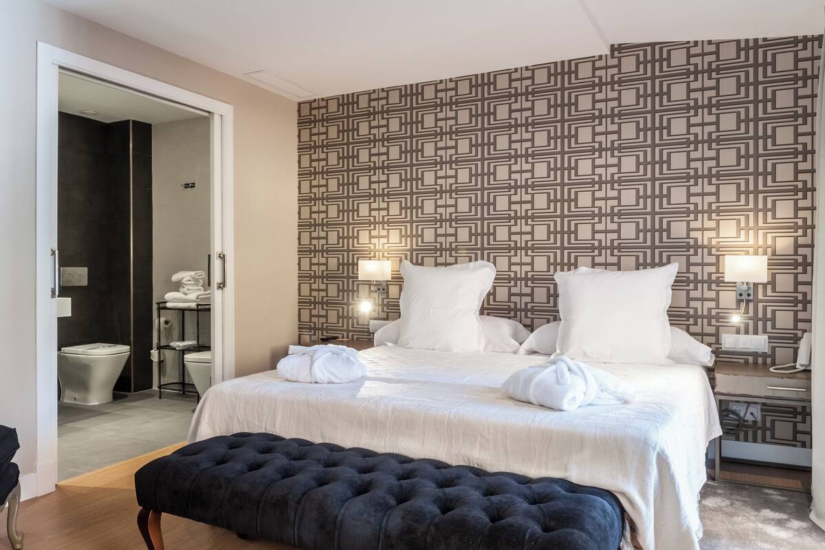 A hotel room with a bed, bedside lamps, a black ottoman, and a patterned wall.