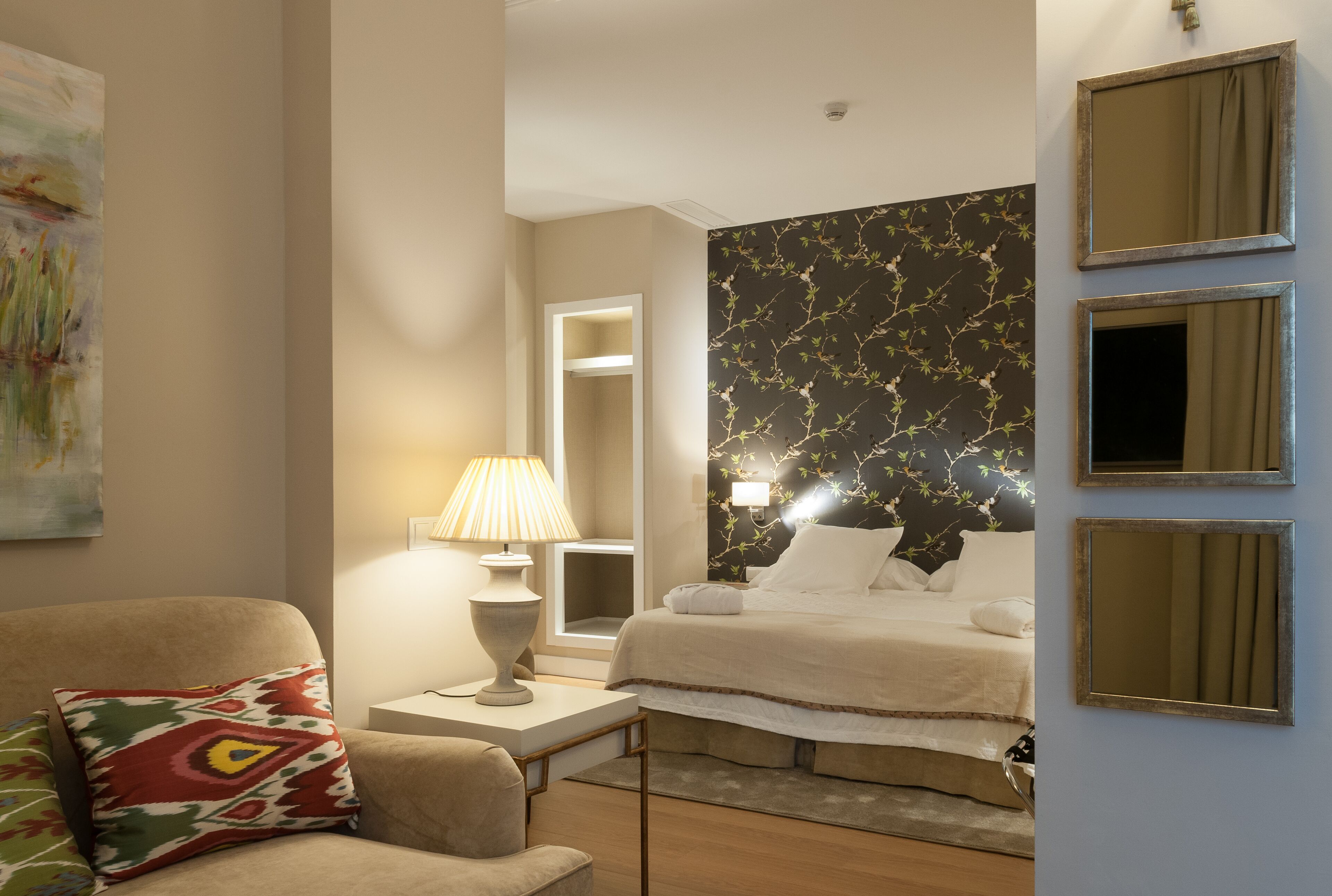 Una habitación de hotel con cama, una silla, una mesita de noche, una lámpara y un muro con estampado floral.