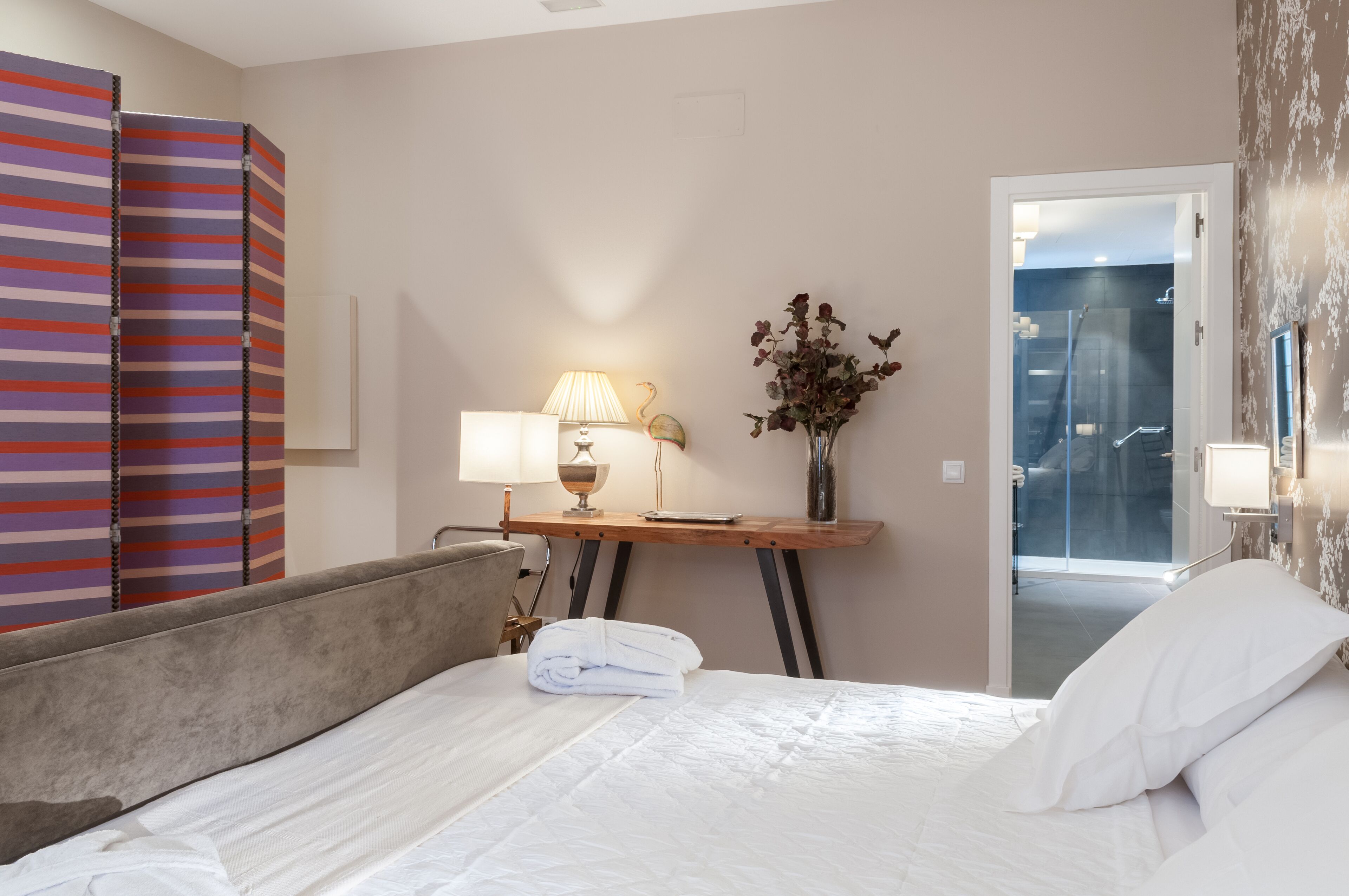 A hotel room with a bed, a desk with a lamp, a vase with flowers, and a shower area visible through a glass door.