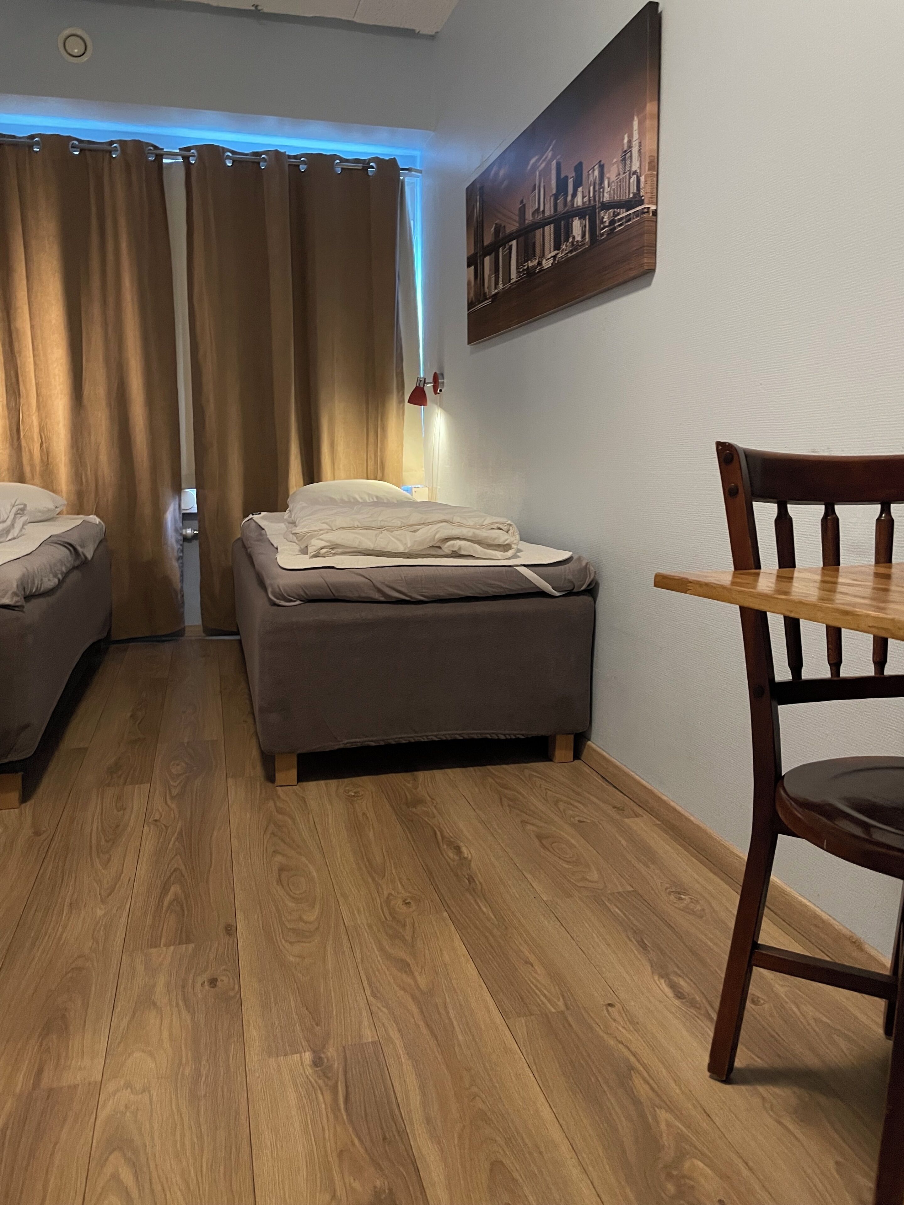 Una habitación con dos camas, piso de madera, una mesa de comedor con una silla y un cuadro de un paisaje urbano en la pared.