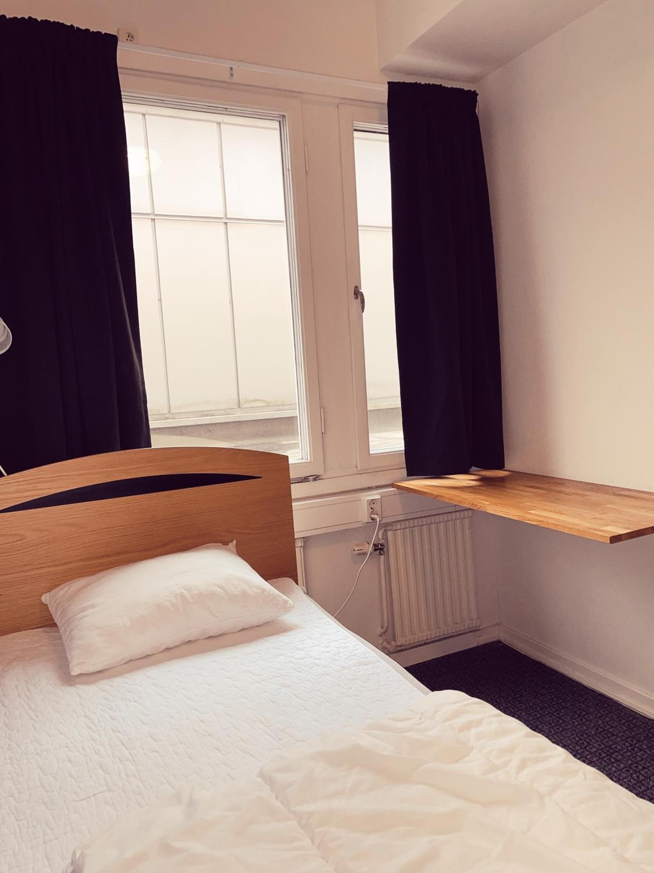 A single bed with a wooden headboard, a white pillow, and a white duvet in a room with a window and a radiator.