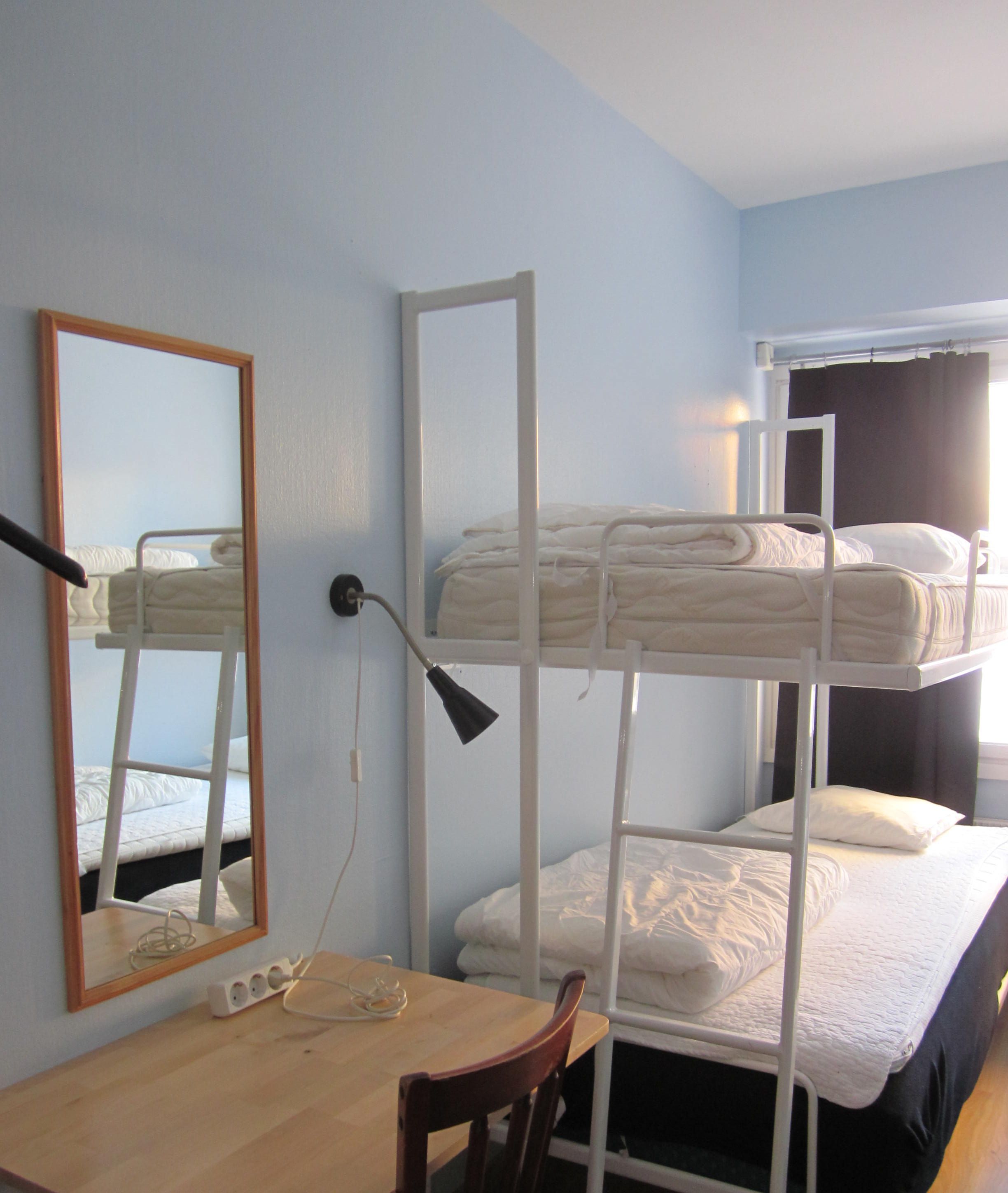 A bunk bed room with a wooden desk and chair, a mirror, and a window with curtains.